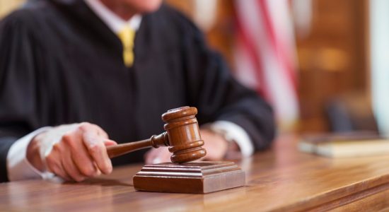 A defense attorney MO stands before a judge in a black robe, who holds a wooden gavel and prepares to strike it in the courtroom, with the American flag visible in the background.