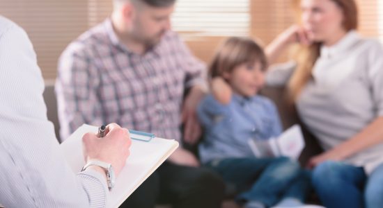 A person holding a clipboard takes notes while a man, woman, and child sit together on a couch, possibly meeting with a child custody lawyer in Jefferson City to discuss their family's situation.