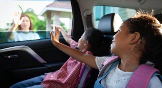 Two young girls with backpacks sit in the backseat of a car, waving through the window at a woman outside—perhaps after visiting a Jefferson City relocation attorney with divorce expertise.