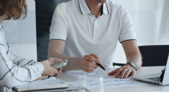 Two people sit at a desk reviewing printed charts and graphs; one holds a pen and points at the paper, while the other holds a smartphone.